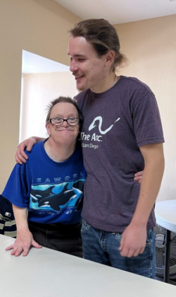 A man smiles as he gives a side hug to another man smiling, with glasses