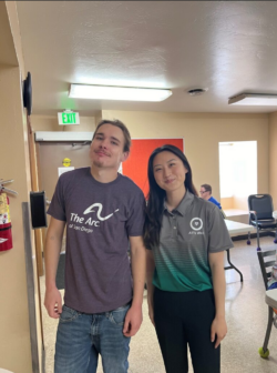 A man and woman smile together, the woman is wearing an All's Well shirt and a man is wearing an Arc tshirt