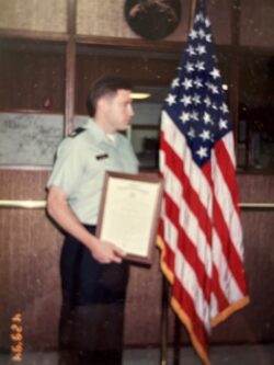 Pat Flynn in a military uniform holding a framed letter and standing next to an American flag