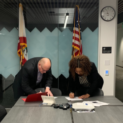 JaRita and Pete sign papers on a long table in a large conference room