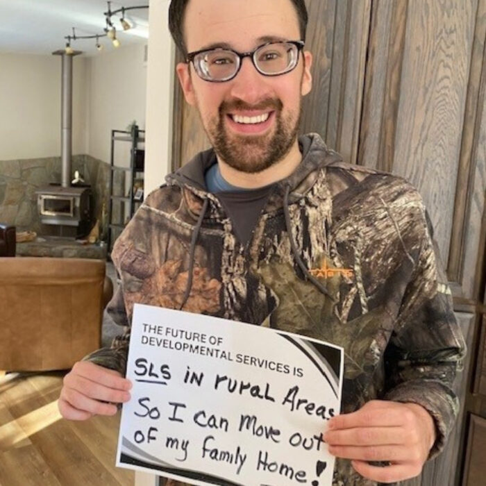 Evan Libby wearing glasses smiles at the camera while holding a sign that reads The Future of Developmental Services is SLS in rural areas so I can move out of my family home