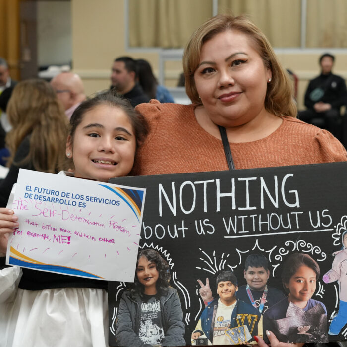 alt="Karina Saldaña smiles at the camera next to a little girl while the little girl a holds sign that read El Futuro de los Servicios de Desarrollo es The Self Determination Program that brings peace and helps others for example me and Karina holds a sign with a collage of kids that include the girl standing next to her and the words Nothing about us without us"