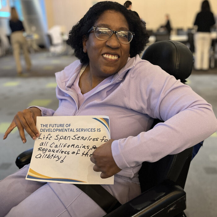 Lisa Cooley smiles at the camera while sitting on a wheelchair and holding a sign that reads The Future of Developmental Services is Life Span services for All Californians Regardless of their Ability