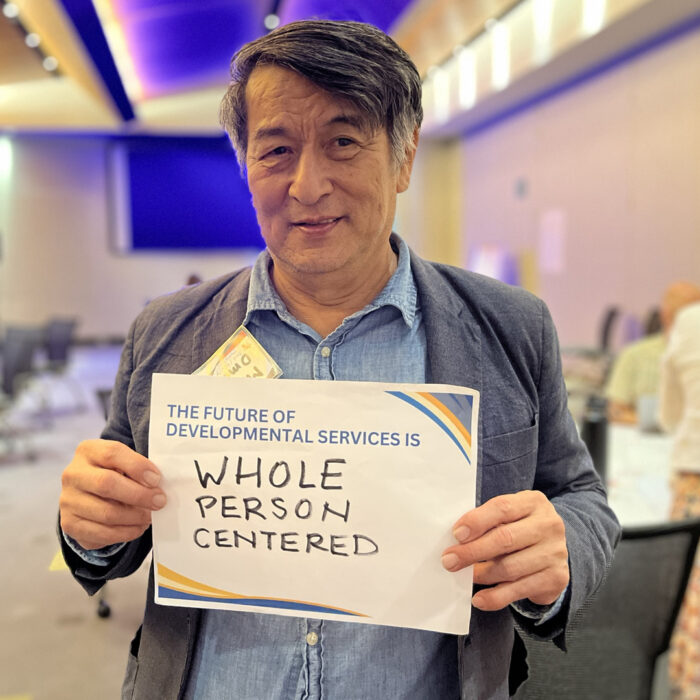 Marty smiles at the camera while holding a sign that reads The Future of Developmental Services is whole person centered