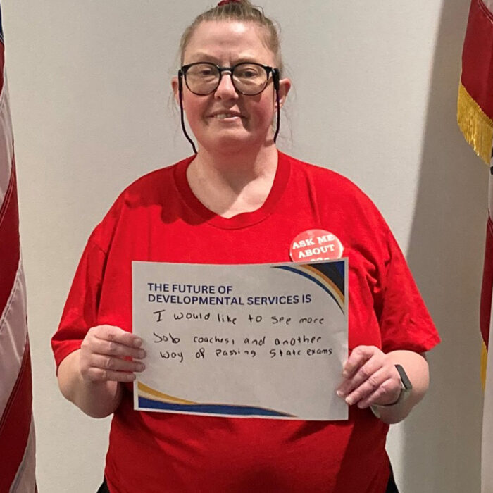 Michelle Padilla wearing glasses smiles at the camera while holding a sign that reads The Future of Developmental Services is I would like to see more job coaches and another way of passing state exams