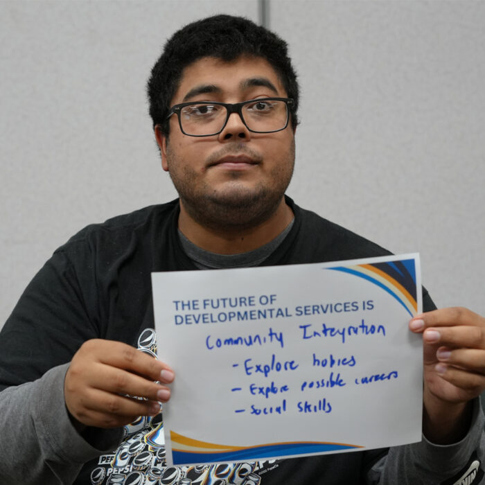 Nestor wearing glasses looks at the camera while holding a sign that reads The Future of Developmental Services is Community Integration explore hobbies explore possible careers social skills