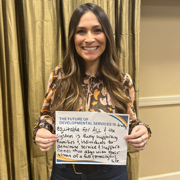 Season smiles at the camera while holding a sign that reads The Future of Developmental Services is truly equitable for all and the system is fully supporting families and individuals to determine service and support needs that align with their vision of a full and meaningful life