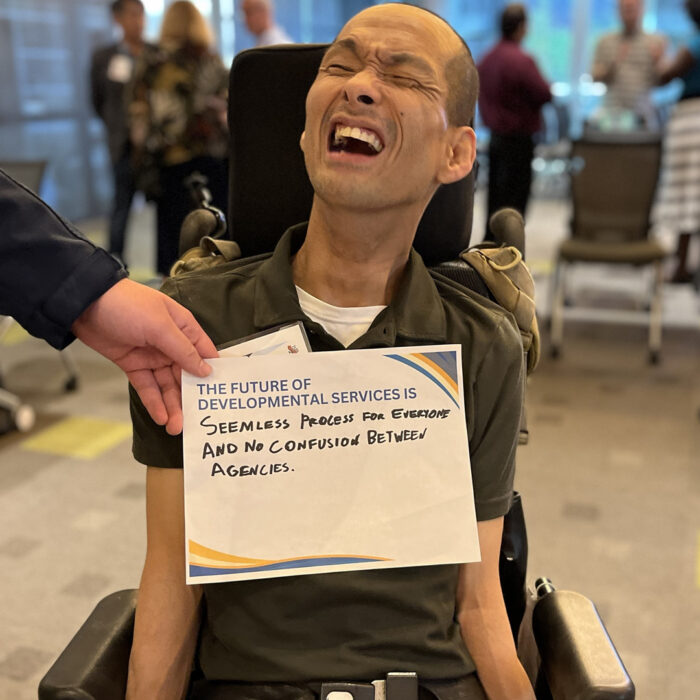 Tim Jin smiles with his eyes closed sitting on a wheelchair while someone a hand of someone off the camera frame holds a sign that reads The Future of Developmental Services is Seamless process for everyone and no confusion between agencies