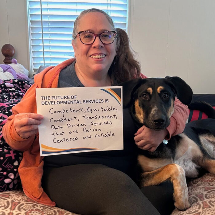 Tina Ewing with glasses sits on a bed embracing her cute dog while and smiles while both look and the camera and she holds a sign that reads The Future of Developmental Services is competent equitable consistent transparent data driven services that are person centered and reliable
