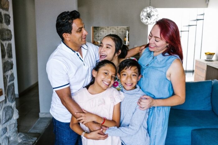 A photo of a Hispanic family in their living room, hugging, smiling and laughing. There is a blue couch and globe chandelier behind them and a stone fireplace to their left. There is a father who is wearing a white polo shirt and jeans. A mother wearing a denim dress. A teenage daughter wearing a floral shirt. A younger daughter wearing a pink dress. A son wearing a grey sweatshirt.