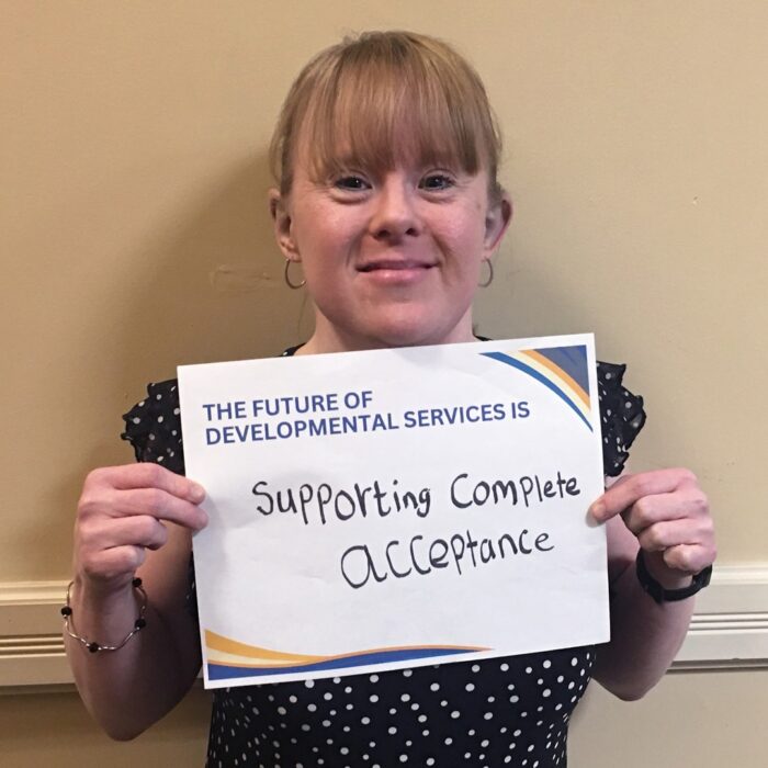 Sara Specks smiles at the camera while holding a sign that reads The Future of Developmental Services is supporting complete acceptance