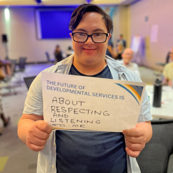 Alexander Taua Brown Omoto smiles at the camera while holding a sign that reads: "The future of developmental services is about respecting and listening to me."