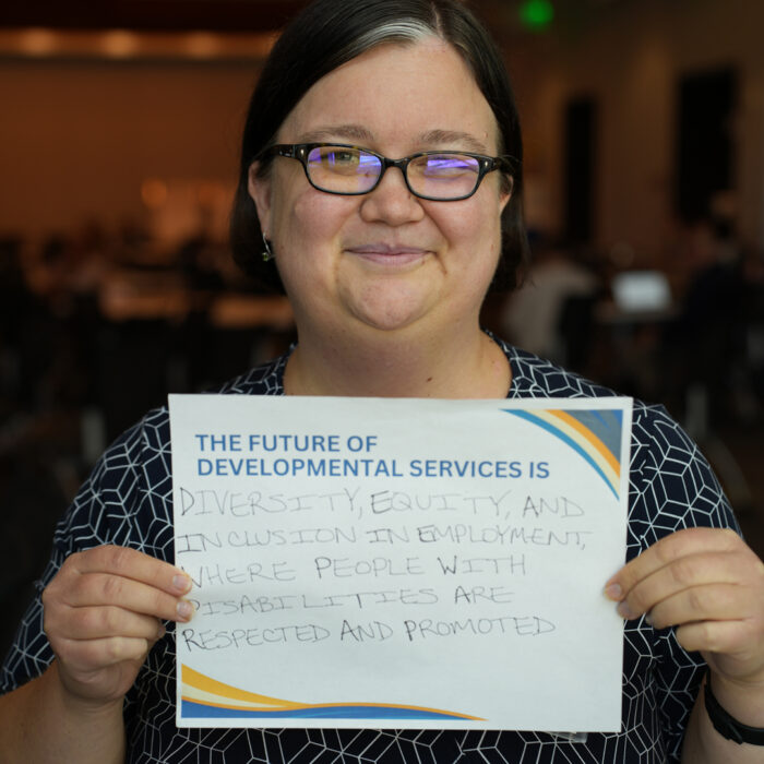 Elena Tiffany smiles at the camera while holding a sign that reads: "The Future of Developmental Services is diversity, equity and inclusion in employment, where people with disabilities are respected and promoted."