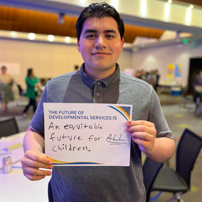 Oscar Mercado smiles at the camera while holding a sign that reads: "The future of developmental services is an equitable future for ALL children."