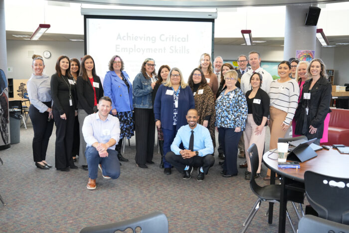 A group of 22 individuals pose for the camera in a room in front of a projector that reads Achieving Critical Employment Skills.