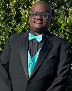 Sean Wilson smiles at the camera while standing in front of some greenery in a black 3-piece suit with teal vest and bow tie.