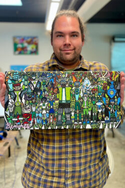 Ben Watts smiles at the camera while holding one of his colorful artworks, which includes many lively characters that appear to be superheroes, robots, sports people, etc.
