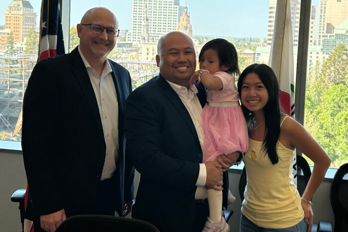 Two men and two girls smile at the camera. One of the men is holding one of the girls who is a toddler. The other girl is standing to the right of that man.