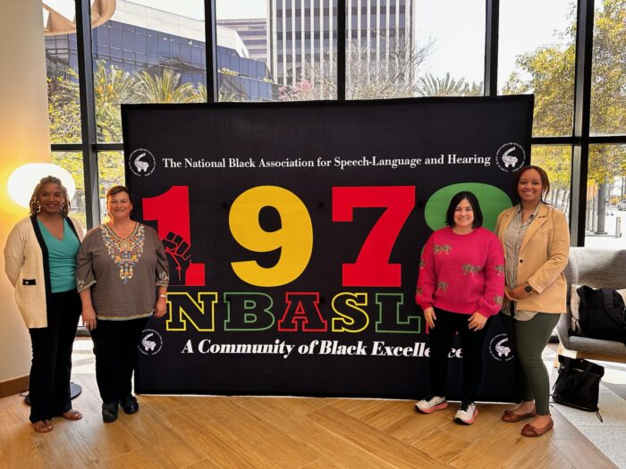 Four smiling women stand in front of a large sign (two on each side of it) inside a building. The sign reads: The National Black Association for Speech-Language and Hearing. 1978 NBASLH. A community of Black Excellence.
