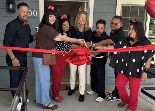 Seven people stand behind a red ribbon on the porch of a house