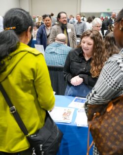 Two women in a busy room face a table with printed materials on it and a blue tablecloth. A smiling woman is talking to them.