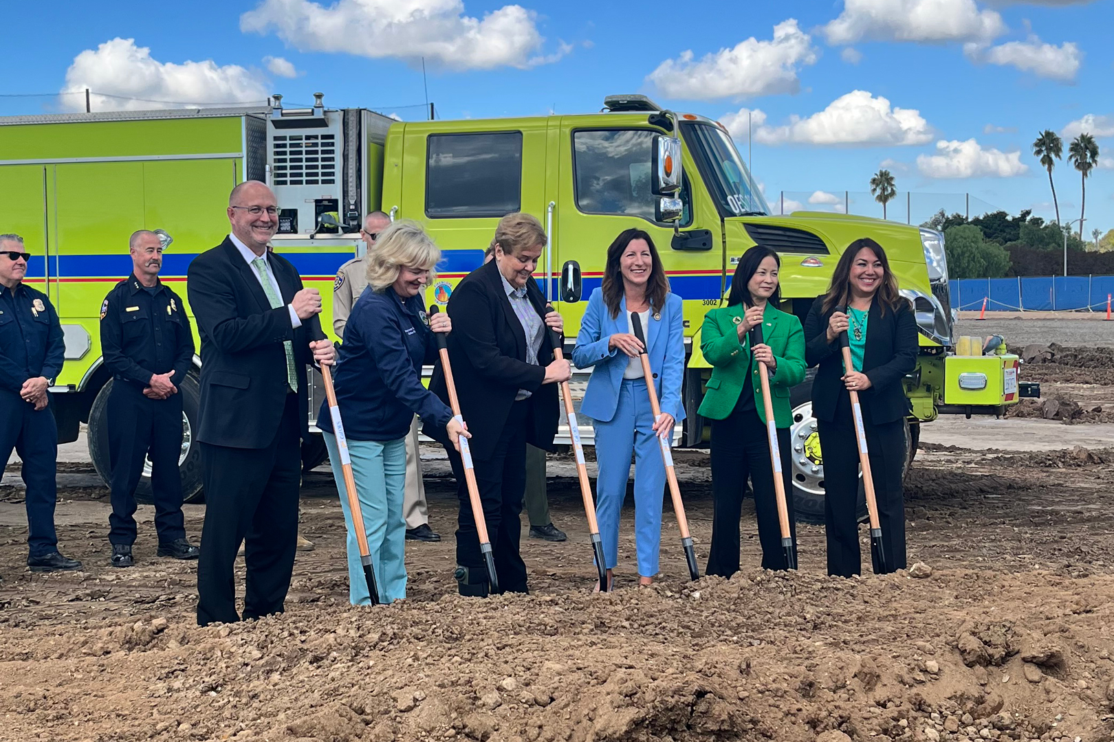 Two men and four women in suits hold shovels in front of a yellow truck.