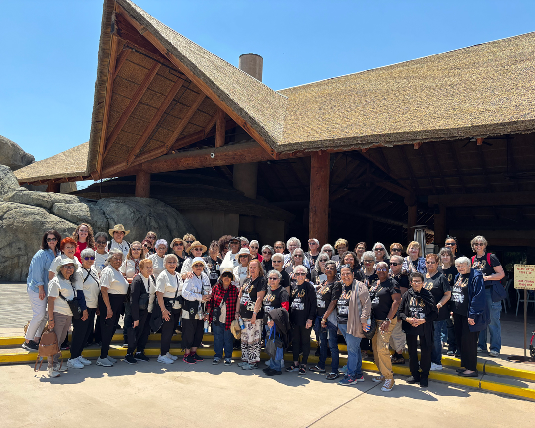 A large group of seniors stand outside of a one-story building with a brown roof.