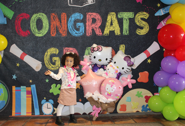 Little girl poses in front of a colorful background that reads congrats and next to a bunch of pink and white ballons in the shape of stars and Hello Kitty.