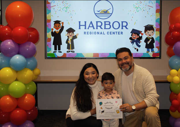 A small boy poses with his mother and father while holding a diploma. In the background there are multi-colored balloons and a screen displaying the Harbor Regional Center logo.Harbor 