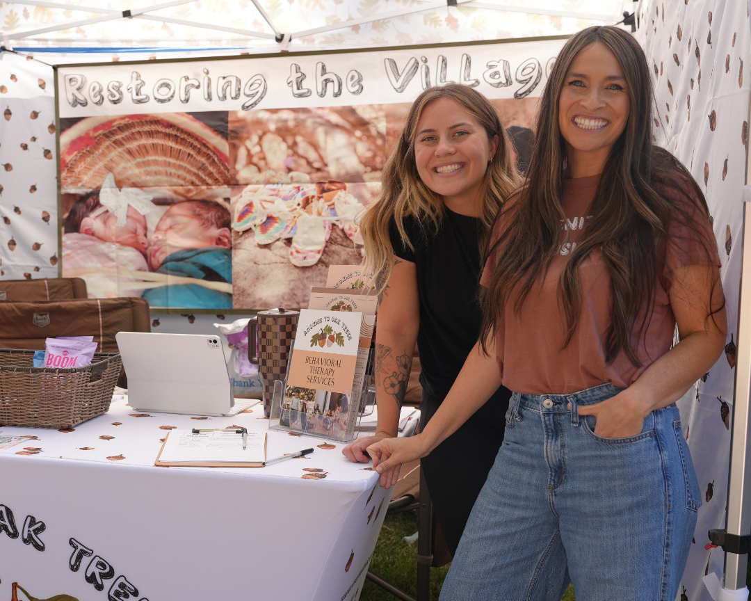 Two women smile at the camera in next to a table with reading materials. There is a banner on the background that reads "Restoring the village."