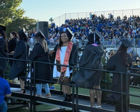 An outdoor graduation ceremony. A woman smiles at the camera as she goes up a ramp dressed in a cap and gown.