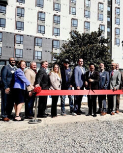 Eleven people in suits stand behind a red ribbon in front of a building.