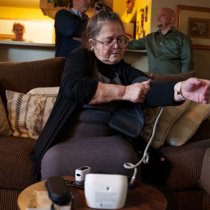 Woman sitting on a couch putting on a blood pressure cuff while three stand in the background